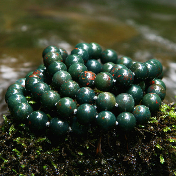 A Bloodstone healing gemstone necklace, lays on green moss near a stream, known for boosting the immune system through crystal healing